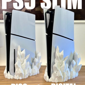 May include: Two white PS5 Slim consoles, one with a disc drive and one without, are shown on a wooden surface. Both consoles are resting on a white, 3D-printed stand that looks like a cluster of crystals.