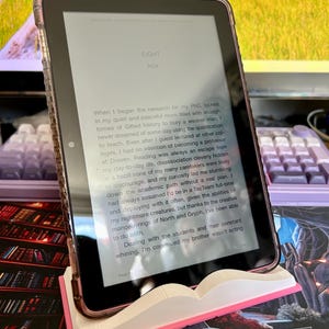 May include: A digital tablet with a clear case, displaying text from a book, resting on a pink and white book-shaped stand. The text on the screen reads "EIGHT" and "NOX".
