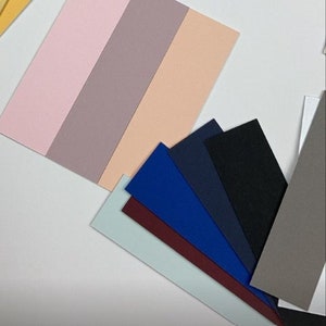 Peut inclure: Un éventail de nuanciers de couleurs dans différentes nuances de rose, beige, bleu, vert et gris. Les échantillons sont disposés en éventail sur une surface blanche. Le texte "Colour sample photoshoot" est écrit en noir sur fond blanc avec une icône d'appareil photo.