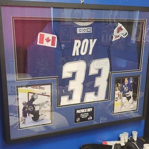 May include: A framed sports memorabilia display featuring a blue Patrick Roy Colorado Avalanche jersey with the number 33, a Canadian flag patch, an Avalanche logo patch, two action photographs of the goalie, and a nameplate reading PATRICK ROY.