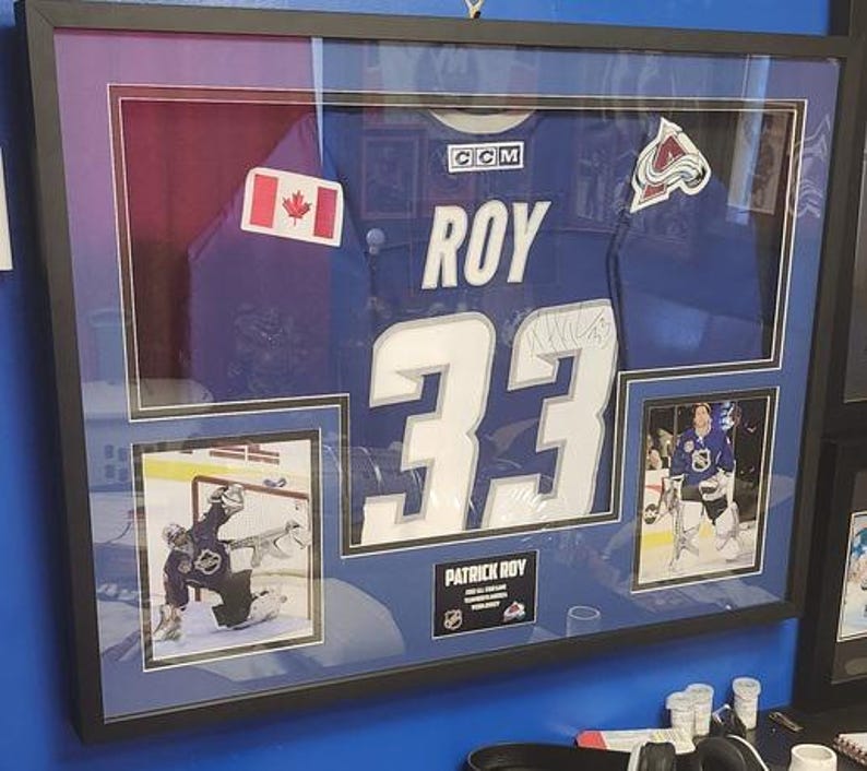 May include: A framed sports memorabilia display featuring a blue Patrick Roy Colorado Avalanche jersey with the number 33, a Canadian flag patch, an Avalanche logo patch, two action photographs of the goalie, and a nameplate reading PATRICK ROY.