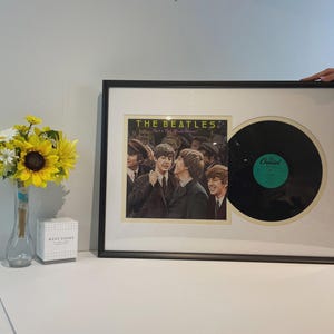 May include: A framed display featuring The Beatles Meet The Beatles album cover art alongside a black vinyl record. The rectangular black frame includes a cream-colored mat board surrounding the square album jacket and the circular record.