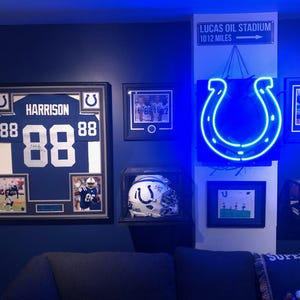 May include: A wall display featuring a framed Indianapolis Colts jersey with the name HARRISON and number 88, a signed helmet in a display case, a glowing blue horseshoe neon sign, a football in a case, and a sign reading LUCAS OIL STADIUM 10 12 MILES.