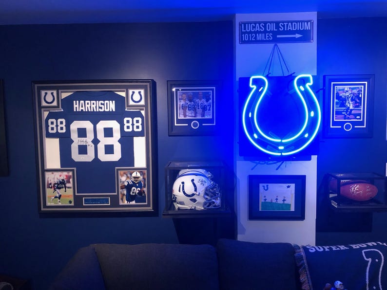 May include: A wall display featuring a framed Indianapolis Colts jersey with the name HARRISON and number 88, a signed helmet in a display case, a glowing blue horseshoe neon sign, a football in a case, and a sign reading LUCAS OIL STADIUM 10 12 MILES.