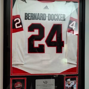 May include: A framed white hockey jersey with red and black accents, featuring the name BERNARD-DOCKER and the number 24 in black. The display includes a small team logo patch, a player photograph, and a descriptive plaque in a black frame.