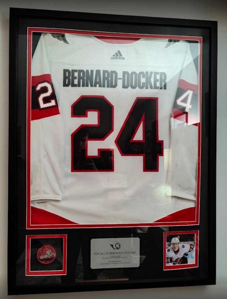 May include: A framed white hockey jersey with red and black accents, featuring the name BERNARD-DOCKER and the number 24 in black. The display includes a small team logo patch, a player photograph, and a descriptive plaque in a black frame.