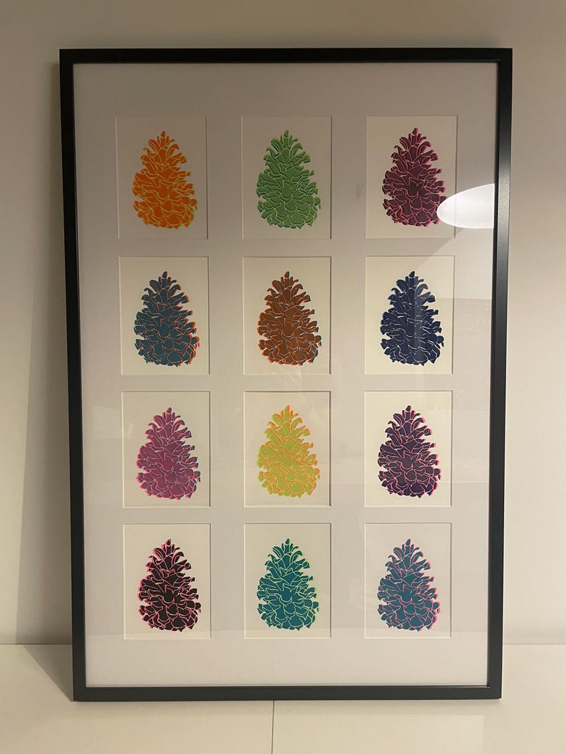 May include: A framed wall art piece featuring a grid of twelve individual prints of pinecones in various vibrant colors including orange, green, magenta, blue, brown, yellow, and teal, all set against a white background within a black frame.