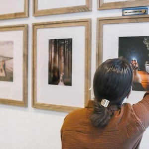 May include: A collection of light wood picture frames with white matting mounted on a white wall. Each frame contains a photograph. A blue leveling tool is positioned on top of one frame to ensure it is straight while being hung on the wall.
