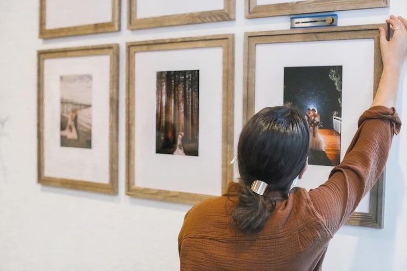 May include: A collection of light wood picture frames with white matting mounted on a white wall. Each frame contains a photograph. A blue leveling tool is positioned on top of one frame to ensure it is straight while being hung on the wall.