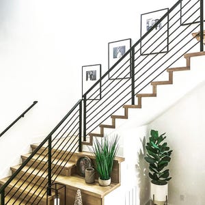 May include: A staircase with light wood treads and a black metal railing featuring horizontal bars. The wall displays three framed black and white photographs. Decorative items include a potted plant, a lantern, and small vessels on the steps.
