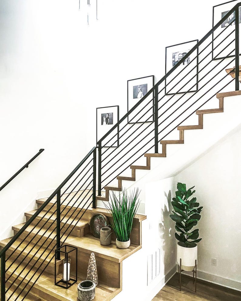 May include: A staircase with light wood treads and a black metal railing featuring horizontal bars. The wall displays three framed black and white photographs. Decorative items include a potted plant, a lantern, and small vessels on the steps.