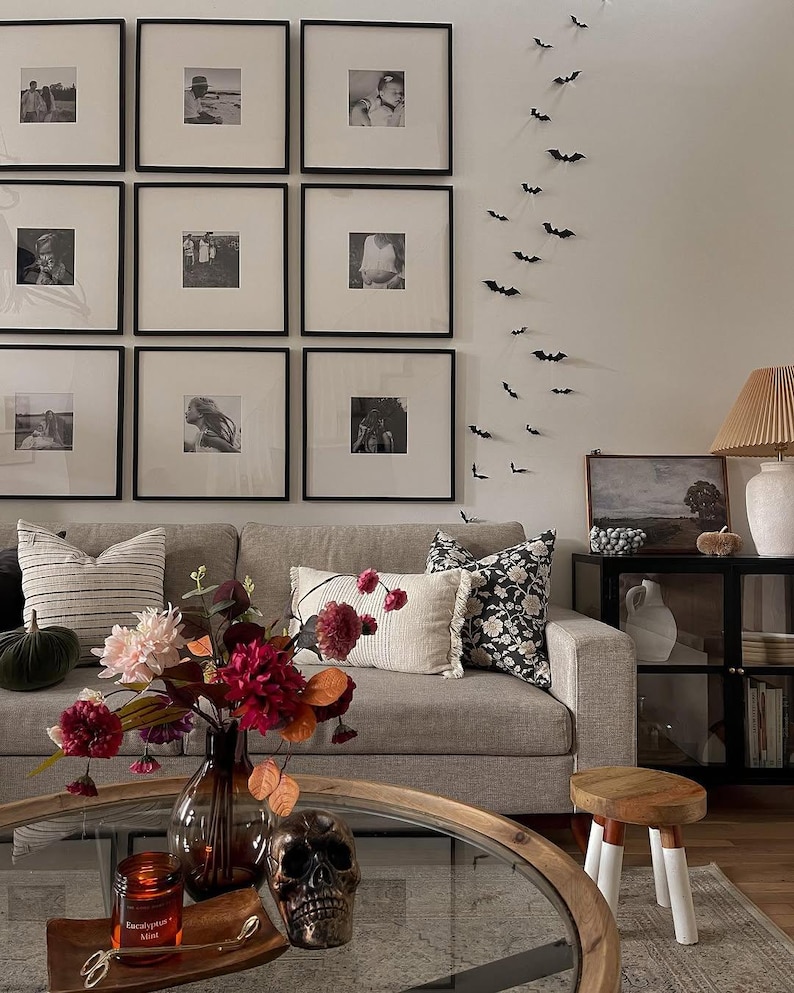 May include: A living room featuring a light gray sofa with striped and floral pillows, a glass coffee table with a floral arrangement, a skull, and a candle, a wall with nine framed black and white photos, and a vertical display of small black bats.