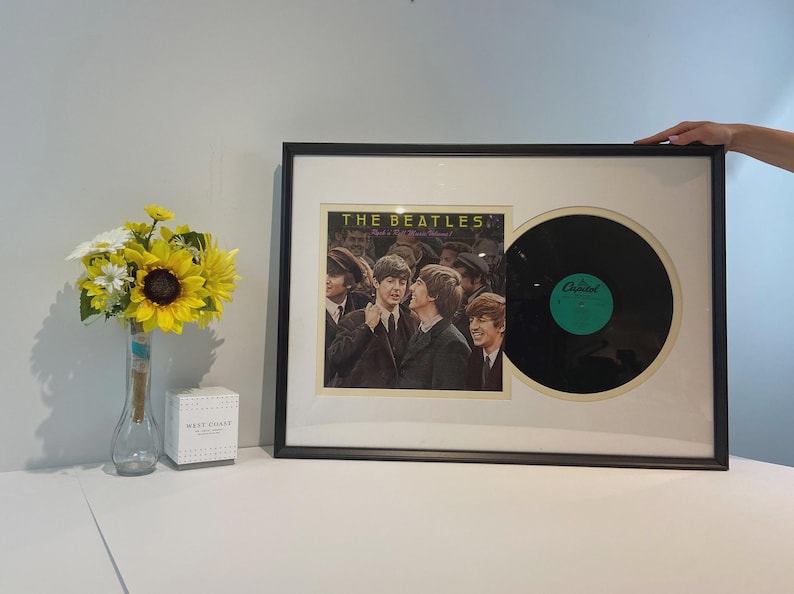 Może przedstawiać: Oprawiona ekspozycja przedstawiająca okładkę albumu The Beatles Meet The Beatles wraz z czarną płytą winylową. Prostokątna czarna ramka zawiera kremowe passe-partout otaczające kwadratową okładkę albumu oraz okrągłą płytę.