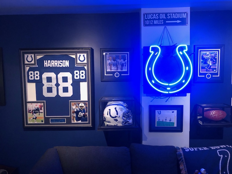 May include: A wall display featuring a framed Indianapolis Colts jersey with the name HARRISON and number 88, a signed helmet in a display case, a glowing blue horseshoe neon sign, a football in a case, and a sign reading LUCAS OIL STADIUM 10 12 MILES.