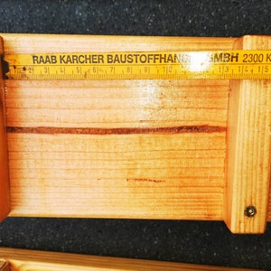 Pu&ograve; includere: Una struttura in legno con un righello pieghevole posizionato sopra. Il righello mostra le misure in centimetri. Il testo "RAAB KARCHER BAUSTOFFHAND" e "MBH 2300 Kiel 14" &egrave; visibile sul righello.