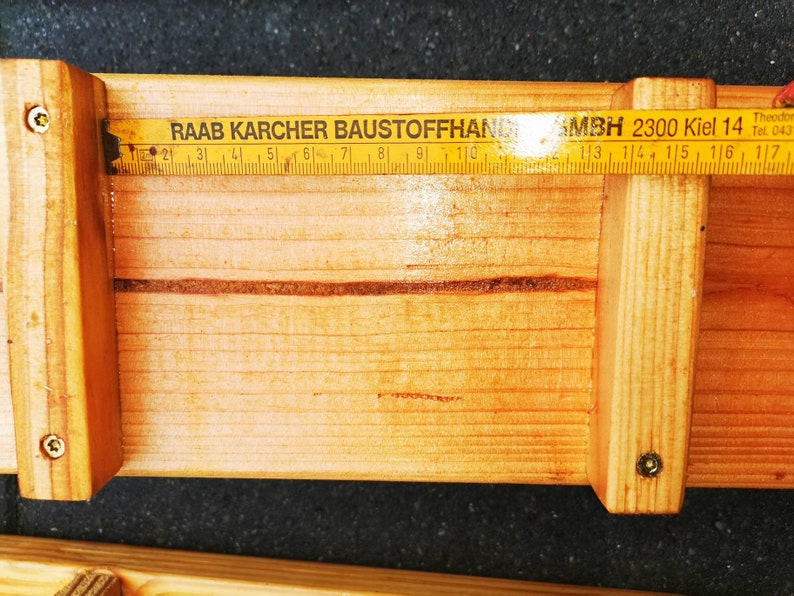 Pu&ograve; includere: Una struttura in legno con un righello pieghevole posizionato sopra. Il righello mostra le misure in centimetri. Il testo "RAAB KARCHER BAUSTOFFHAND" e "MBH 2300 Kiel 14" &egrave; visibile sul righello.