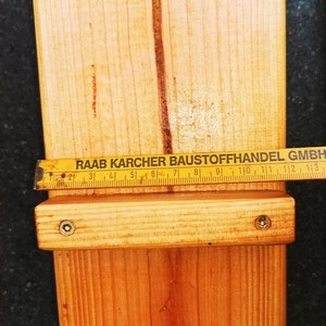 Pu&ograve; includere: Primo piano di assi di legno con un metro a nastro giallo. Il nastro, segnato in centimetri, indica "RAAB KARCHER BAUSTOFFHANDEL GMBH 2300 Kiel". Le assi sono unite con viti. Falegnameria e costruzione.