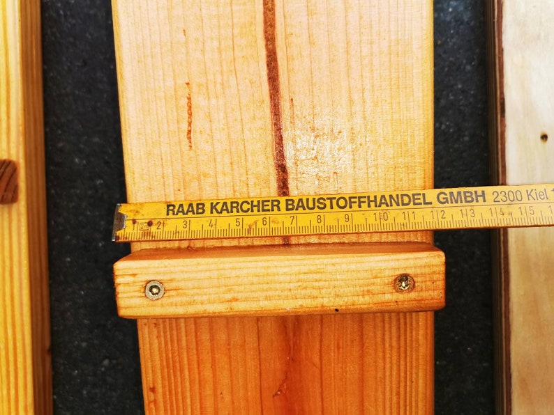 Pu&ograve; includere: Primo piano di assi di legno con un metro a nastro giallo. Il nastro, segnato in centimetri, indica "RAAB KARCHER BAUSTOFFHANDEL GMBH 2300 Kiel". Le assi sono unite con viti. Falegnameria e costruzione.