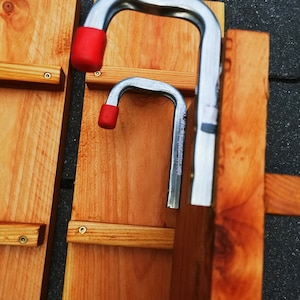 Pu&ograve; includere: Primo piano di due assi di legno con staffe metalliche. Le staffe sono argentate con punte in gomma rossa. Le assi sono di colore marrone chiaro e sono fissate con viti. Lo sfondo &egrave; una superficie grigio scuro.