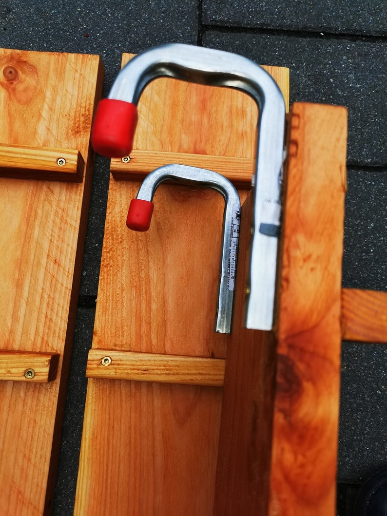 Pu&ograve; includere: Primo piano di due assi di legno con staffe metalliche. Le staffe sono argentate con punte in gomma rossa. Le assi sono di colore marrone chiaro e sono fissate con viti. Lo sfondo &egrave; una superficie grigio scuro.