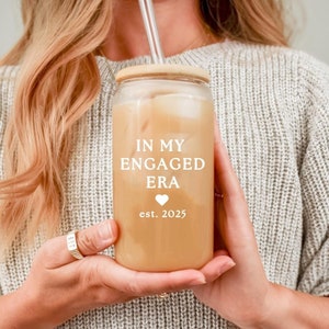 May include: A clear glass tumbler with a wooden lid and a straw. The tumbler is filled with a brown liquid and has the text "IN MY ENGAGED ERA est. 2025" printed on it.
