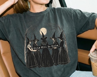 May include: A charcoal gray short-sleeve t-shirt featuring a graphic print of five witches in long dresses and pointed hats standing in a circle under a full moon. The text Girls Will Be Girls is printed in white across the center of the illustration.