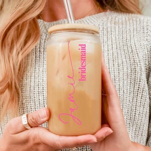 Puede incluir: Un vaso de vidrio esmerilado con tapa de bamb&uacute; y pajita transparente, lleno de una bebida helada. El vaso presenta el nombre Jamie en una tipograf&iacute;a cursiva rosa brillante, con la palabra bridesmaid impresa en una fuente rosa m&aacute;s peque&ntilde;a.