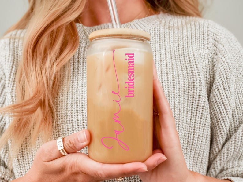 Puede incluir: Un vaso de vidrio esmerilado con tapa de bamb&uacute; y pajita transparente, lleno de una bebida helada. El vaso presenta el nombre Jamie en una tipograf&iacute;a cursiva rosa brillante, con la palabra bridesmaid impresa en una fuente rosa m&aacute;s peque&ntilde;a.
