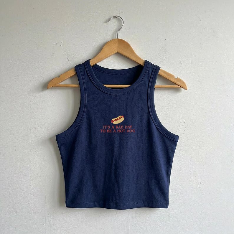 以下が含まれることがあります： ネイビーブルーのリブ編みタンクトップ。ホットドッグの刺繍と「IT'S A BAD DAY TO BE A HOT DOG」という赤い文字が施されています。袖なしのラウンドネックで、木製のハンガーに掛けられています。