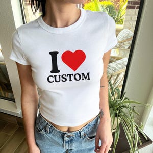 May include: A white short-sleeved crew neck t-shirt featuring the black text I and CUSTOM with a red heart symbol in between. The shirt is worn with blue denim jeans. The design is centered on the chest of the fitted cotton garment.