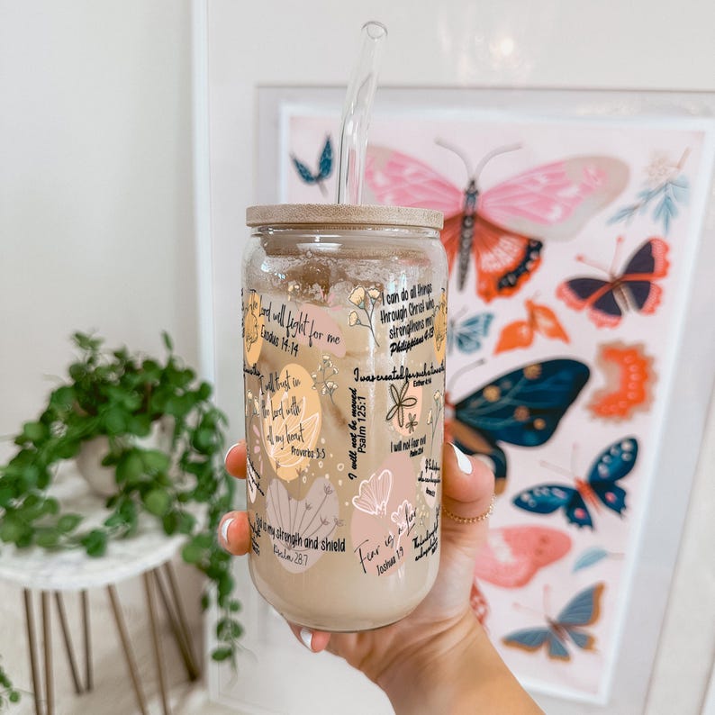 May include: A clear glass tumbler with a wooden lid and a glass straw. The tumbler is decorated with floral designs and inspirational text. The drink inside is light brown. A framed butterfly print is in the background.