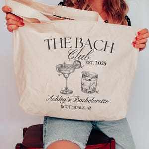 May include: A cream-colored canvas tote bag featuring black printed text that reads THE BACH CLUB EST. 2025, Ashley's Bachelorette, and SCOTTSDALE, AZ. The design includes black line art illustrations of a margarita glass and a rocks glass.