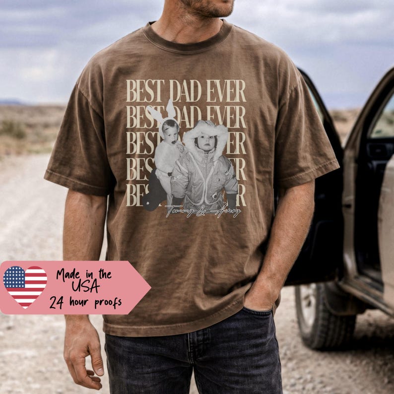 May include: Brown t-shirt with the text "BEST DAD EVER" in a stacked design, featuring a sepia-toned photo of a child. The shirt is worn by a person standing next to a vehicle. The shirt also has the text "Made in the USA" and "24 hour proofs".