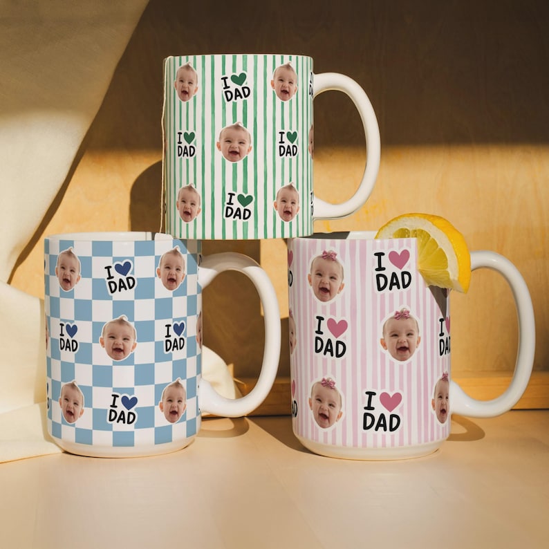 K&ouml;nnte beinhalten: Drei Keramikbecher mit wei&szlig;en Henkeln. Ein Becher hat ein blau-wei&szlig;es Karomuster, ein anderer gr&uuml;ne Streifen und der dritte rosa Streifen. Jeder Becher zeigt Babygesichter und den Text "I ❤️ DAD."