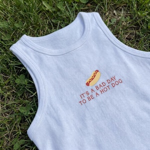 以下が含まれることがあります： ホットドッグの刺繍グラフィックと「IT'S A BAD DAY TO BE A HOT DOG」という赤い文字が施された、白いリブ編みのタンクトップ。丸首で袖のないデザインで、シンプルで清潔感のある外観が特徴です。