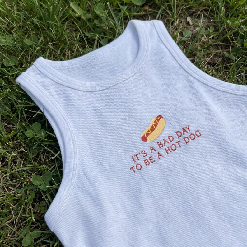 以下が含まれることがあります： ホットドッグの刺繍グラフィックと「IT'S A BAD DAY TO BE A HOT DOG」という赤い文字が施された、白いリブ編みのタンクトップ。丸首で袖のないデザインで、シンプルで清潔感のある外観が特徴です。