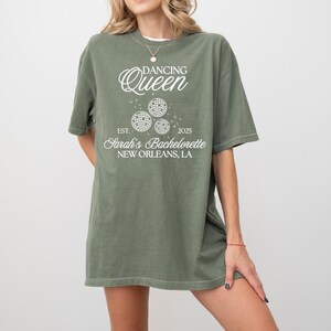 K&ouml;nnte beinhalten: Olivgr&uuml;nes T-Shirt mit dem Text "Dancing Queen" &uuml;ber drei Discokugel-Grafiken. Darunter steht "Est. 2025 Sarah's Bachelorette New Orleans, LA". Das Shirt hat kurze &Auml;rmel und einen Rundhalsausschnitt.