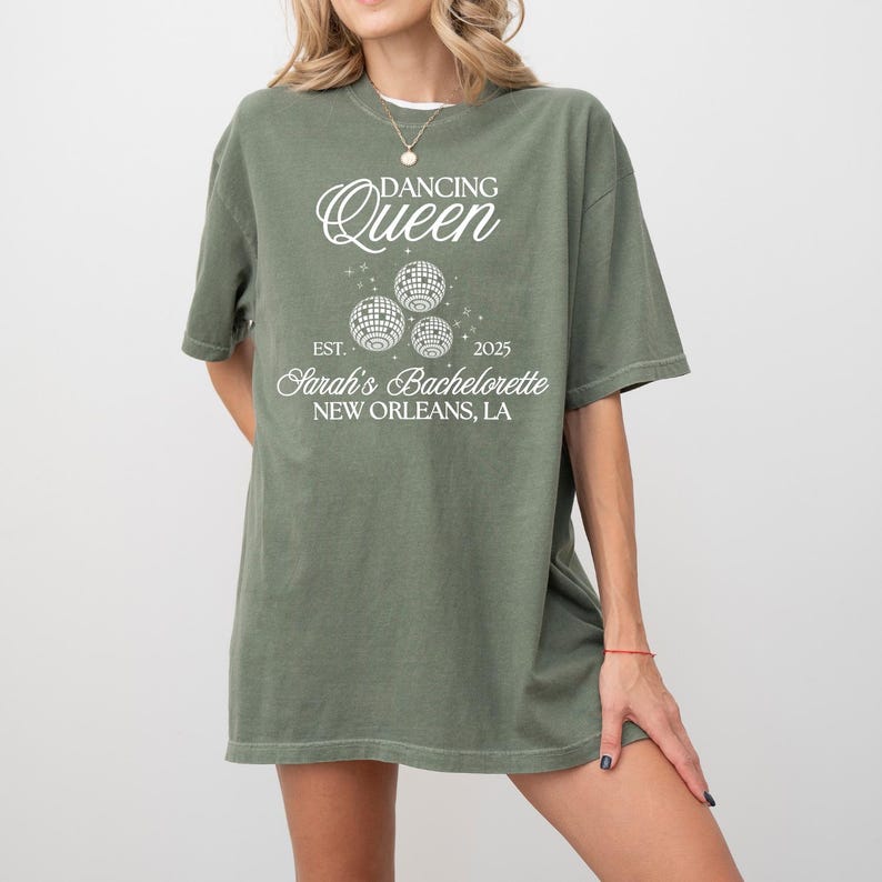 K&ouml;nnte beinhalten: Olivgr&uuml;nes T-Shirt mit dem Text "Dancing Queen" &uuml;ber drei Discokugel-Grafiken. Darunter steht "Est. 2025 Sarah's Bachelorette New Orleans, LA". Das Shirt hat kurze &Auml;rmel und einen Rundhalsausschnitt.