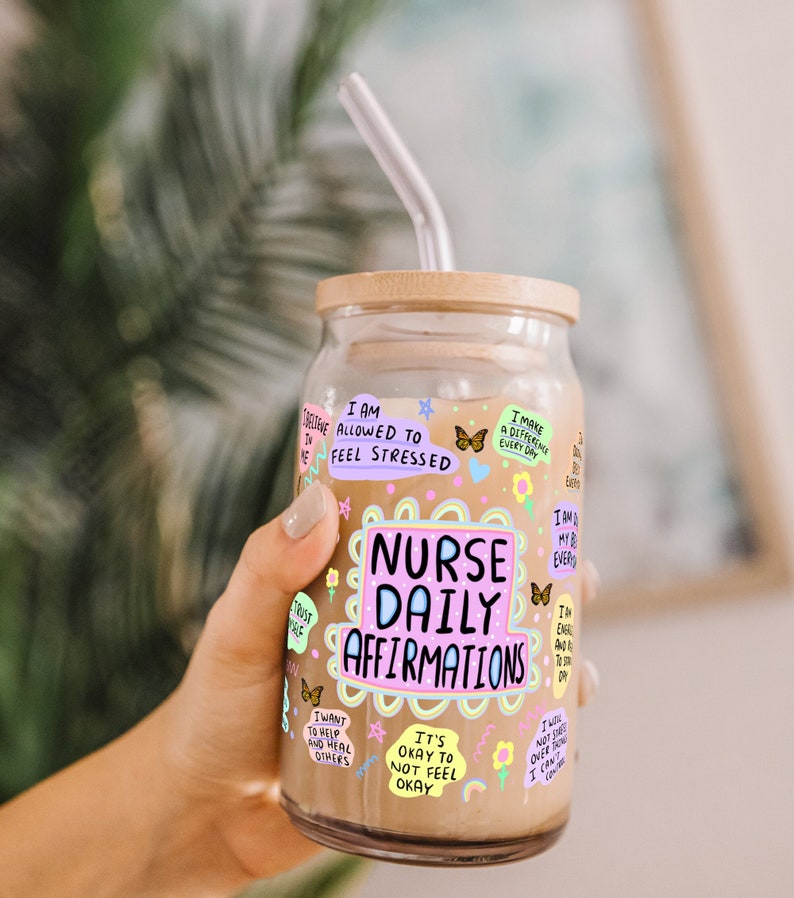 K&ouml;nnte beinhalten: Ein Glasbecher mit Bambusdeckel und einem durchsichtigen Strohhalm. Der Becher ist mit farbenfrohem Text und Illustrationen verziert, darunter die Worte "Nurse Daily Affirmations". Der Becher ist mit einer hellbraunen Fl&uuml;ssigkeit gef&uuml;llt.
