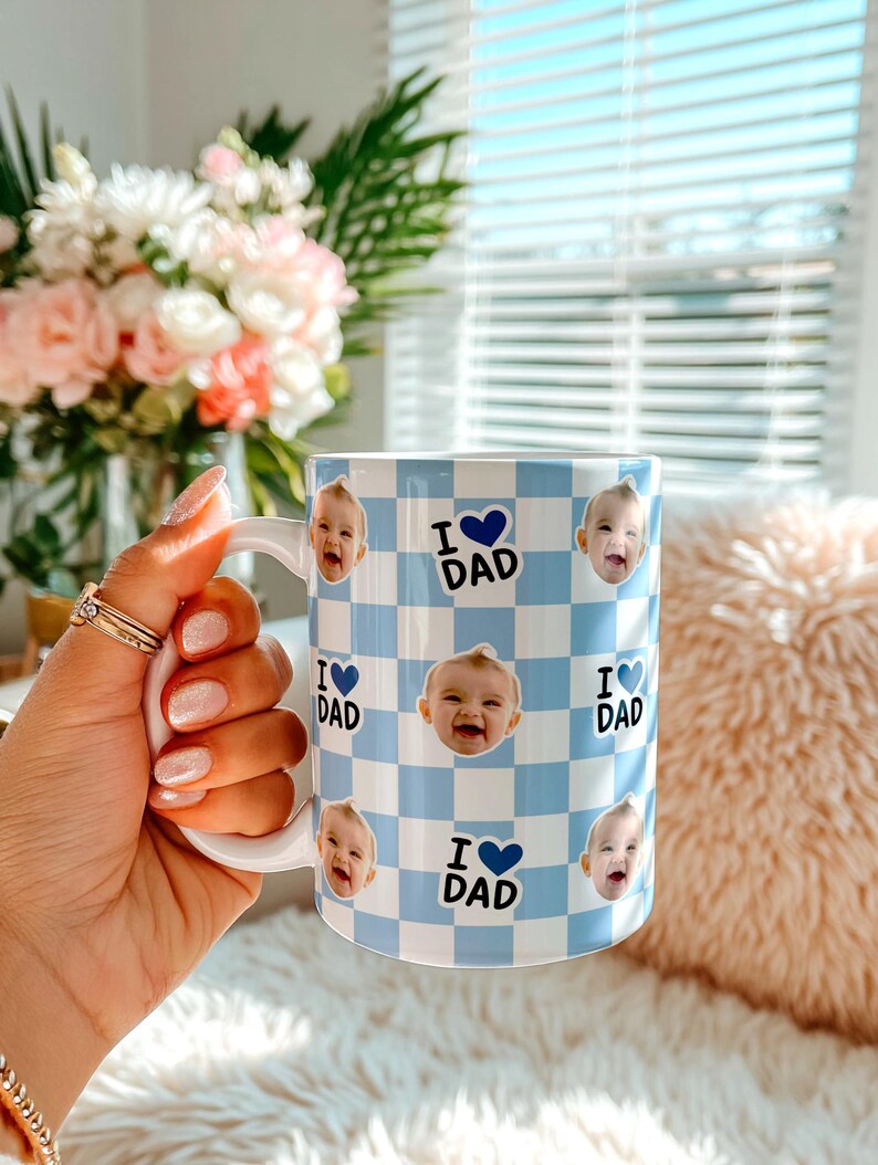 K&ouml;nnte beinhalten: Wei&szlig;e Keramik-Tasse mit blau-wei&szlig;em Karomuster. Die Tasse zeigt Babyfotos und den Text "I ❤️ DAD" in Blau. Die Tasse hat einen wei&szlig;en Henkel.