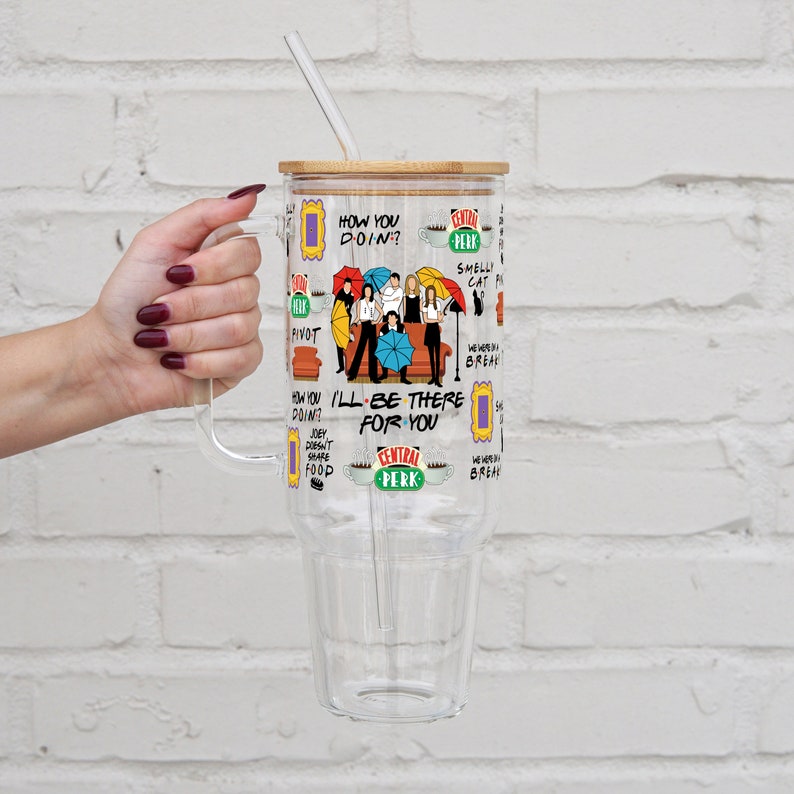 May include: A clear, large tumbler with a bamboo lid and a straw. The tumbler features a colorful graphic with text and images from the TV show "Friends". The handle is clear and the tumbler is held by a hand.