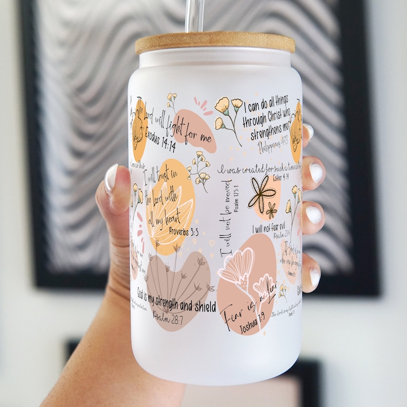 May include: A white glass tumbler with a bamboo lid and a clear straw. The tumbler is decorated with floral designs and inspirational quotes in black script. The quotes include "I can do all things through Christ" and "Fear is a liar."