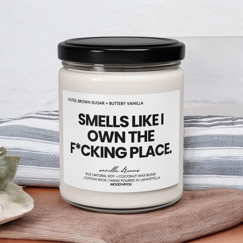 May include: A glass jar candle with a black lid. The label reads "Smells Like I Own The F*cking Place." The candle is vanilla-scented, made with natural soy and coconut wax. The jar is on a brown surface with a striped cloth in the background.