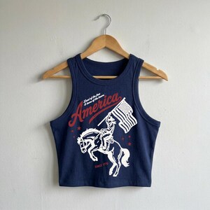 May include: A navy blue ribbed crop tank top featuring a white and red graphic print of a person riding a bucking horse while holding an American flag. The text reads "America", "Land of the free & home of the brave", and "SINCE 1776".