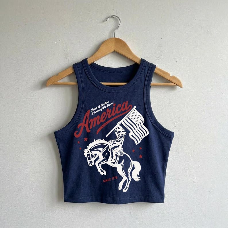 May include: A navy blue ribbed crop tank top featuring a white and red graphic print of a person riding a bucking horse while holding an American flag. The text reads "America", "Land of the free & home of the brave", and "SINCE 1776".