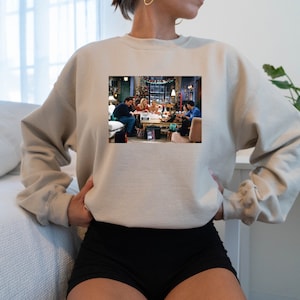 May include: A beige sweatshirt with a printed image of the cast of the TV show "Friends" gathered around a table. The image shows the characters in a living room setting, with Christmas decorations visible in the background.