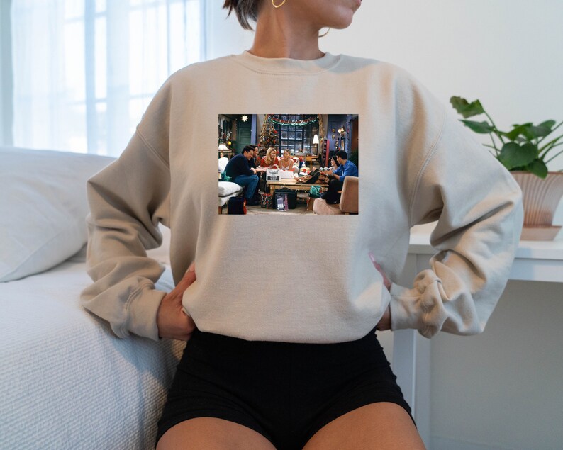 May include: A beige sweatshirt with a printed image of the cast of the TV show "Friends" gathered around a table. The image shows the characters in a living room setting, with Christmas decorations visible in the background.