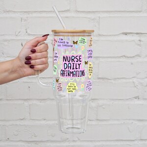 K&ouml;nnte beinhalten: Ein durchsichtiger Glasbecher mit Griff, Bambusdeckel und Strohhalm. Der Becher ist mit bunten Aufklebern und dem Text "NURSE DAILY AFFIRMATIONS" verziert. Der Becher wird von einer Person gehalten.