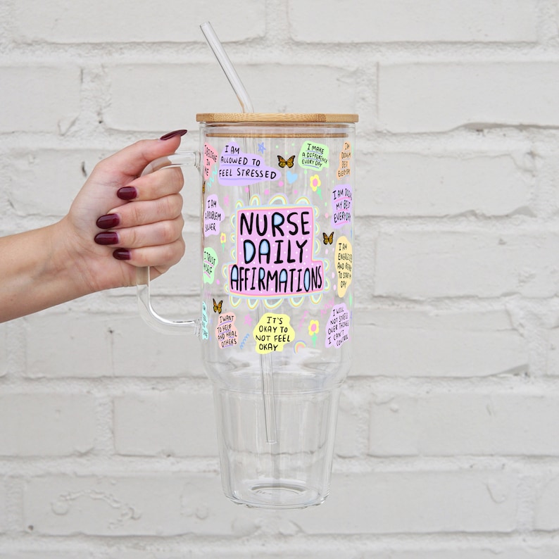 K&ouml;nnte beinhalten: Ein durchsichtiger Glasbecher mit Griff, Bambusdeckel und Strohhalm. Der Becher ist mit bunten Aufklebern und dem Text "NURSE DAILY AFFIRMATIONS" verziert. Der Becher wird von einer Person gehalten.