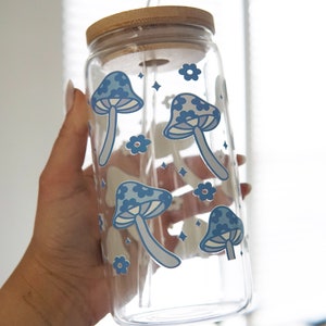 May include: A clear glass tumbler with a bamboo lid and a glass straw. The exterior of the glass features a repeating pattern of blue mushrooms and small flowers, accented with tiny four-pointed star shapes scattered across the surface.